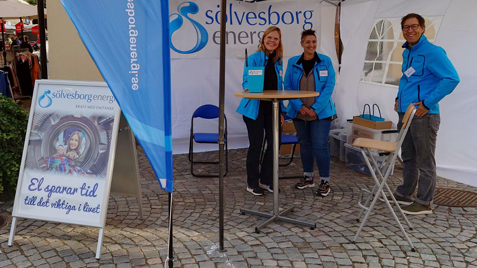 Tre leende personer i ett tält i stadsmiljö sommartid. På flera ställen i bilden syns Sölvesborg Energis logotyp, till exempel på en affisch i en gatupratare vid sidan av tältet.