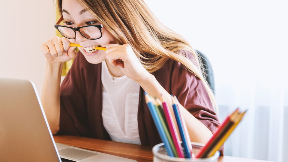 En person med uppspärrade ögon sitter framför en laptop och biter i en penna för att koncentrera sig bättre. Framför henne står ett pennställ.