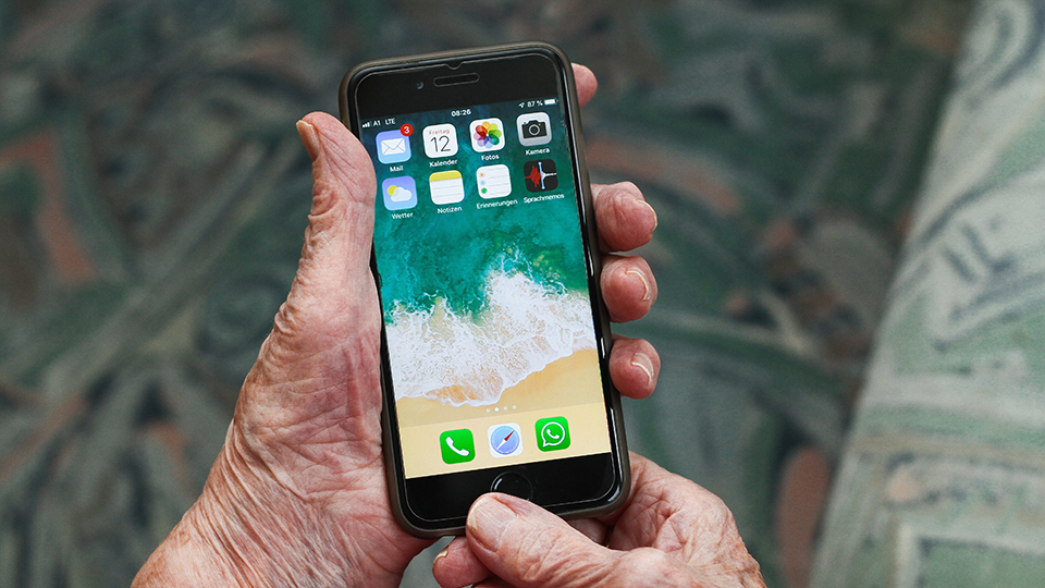 En äldre persons hand håller i en smartphone, på startskärmen finns appar som till exempel mejl, kalender, kamera, väder, anteckningar.