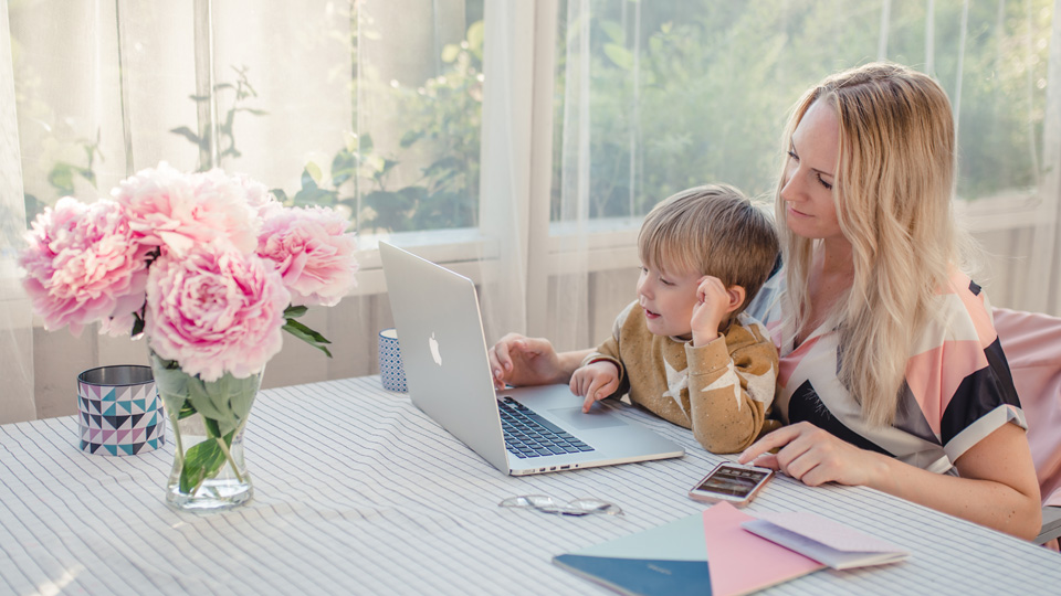 Ett barn och en förälder sitter vid datorn i/på vad som ser ut att vara ett uterum eller en balkong. Det är sommar och de har blommor i en vas framför sig.