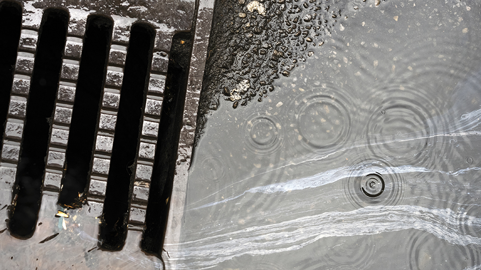 En vattenpöl på asfalt rinner ner i en dagvattenbrunn. På vattenpölens blanka yta blir varje regndroppe flera ringar.